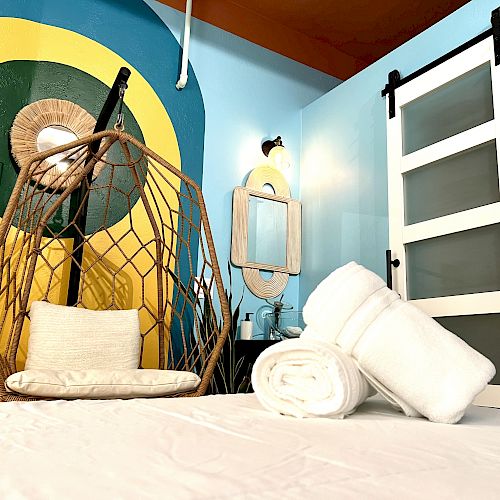 The image shows a modern room with a hanging chair, colorful wall, a mirror, and a set of rolled white towels on a bed.