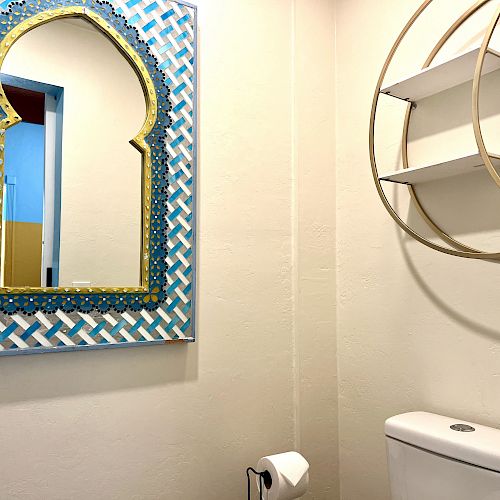 A bathroom with a decorative mirror, a toilet, a toilet paper holder, and a circular wall shelf on a white wall.