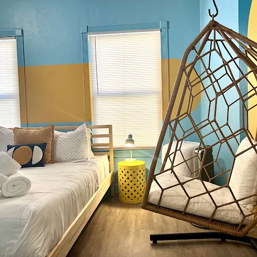 A cozy bedroom with a modern bed, rolled towels, a yellow side table, and a wicker hanging chair, set against blue and yellow walls.