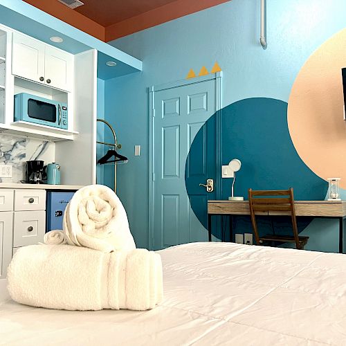 A modern, well-lit room is shown with a bed, kitchen unit, microwave, desk, TV, and neatly rolled towels on the bed, featuring colorful wall decor.