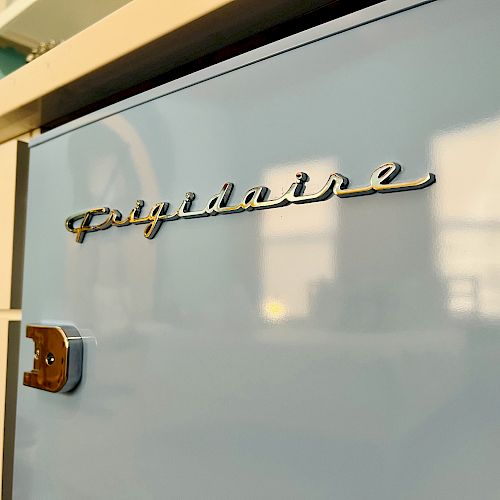 The image shows a close-up of a light blue Frigidaire appliance, with the brand name clearly visible, alongside white cabinetry with metal handles.