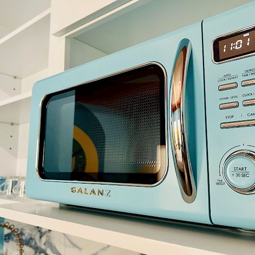 A light blue microwave with a digital clock displaying 11:01 and several buttons on its interface sits on a white shelf, with a bowl beside it.