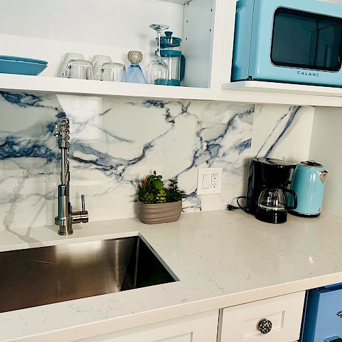 A kitchen setup with a sink, coffee machine, microwave, dish soap, sponge, and potted plant against a marbled backsplash and white countertop.