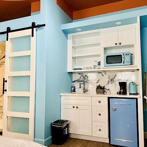 A compact kitchenette with light blue walls has a retro fridge, microwave, coffee maker, and sink. A sliding door leads to a marble-tiled bathroom.