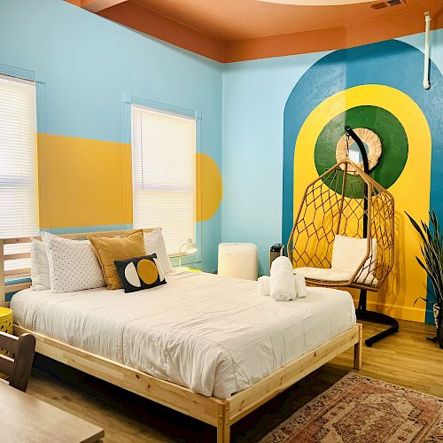 A cozy bedroom with a bed, colorful wall art, windows, blinds, a hanging chair, a rug and plants. The room features vibrant blue, yellow, and green tones.