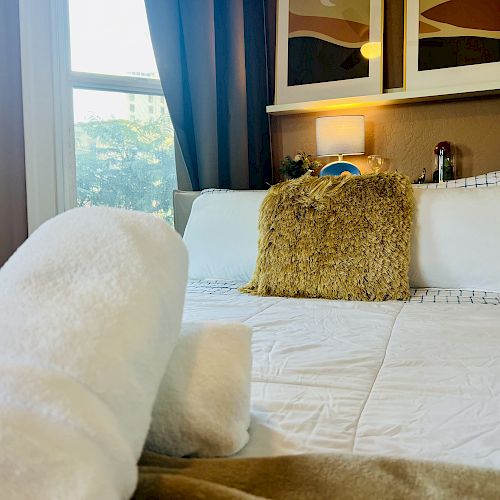 A cozy bedroom with a neatly made bed, a furry throw pillow, wall art above the headboard, and rolled towels on the foreground.