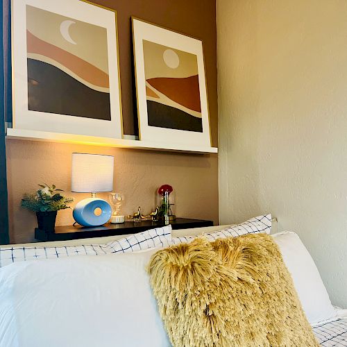 A cozy bed with a fluffy mustard pillow, patterned sheets, two abstract art pieces above, and a nightstand holding a lamp, decor, and a small plant.