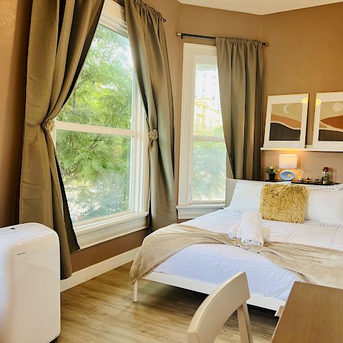 A cozy bedroom with a small bed, desk, chair, large windows, brown curtains, two wall pictures, and a portable air conditioner.