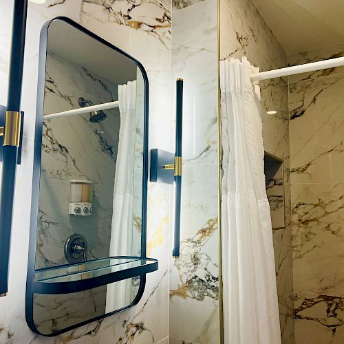 A modern bathroom with marble walls, a large mirror with vertical lights, and a shower area with a white curtain.