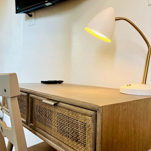 The image shows a wooden desk with drawers, a white chair, and a modern white desk lamp that is turned on. A remote control is on the desk.