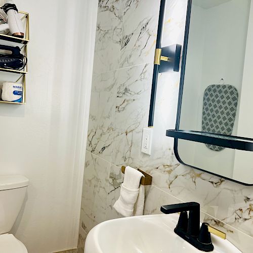 This image shows a bathroom with a single sink, a toilet, and a wall-mounted shelf with towels and toiletries. The wall is tiled.