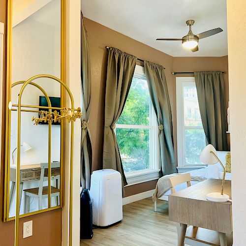 A room with large windows, a ceiling fan, desk, chair, lamp, and an air purifier, framed by brown curtains. A mirror and coat rack are visible on the left.