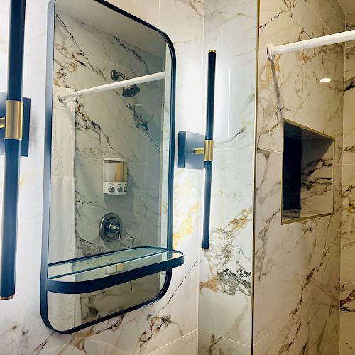 The image shows a modern bathroom with marble walls, a lighted mirror, and a shower area. There's a glass shelf and a built-in soap dispenser.