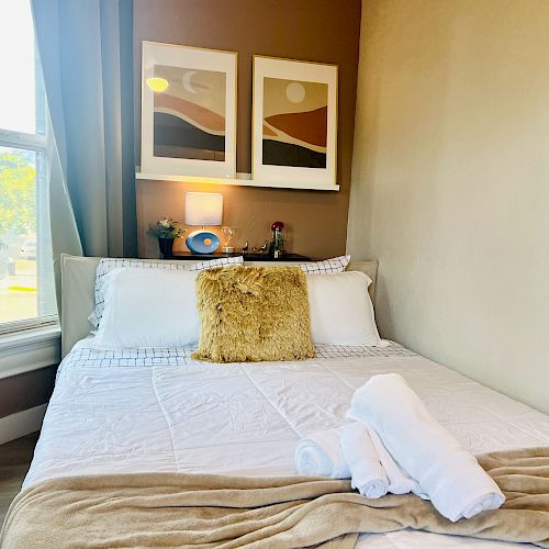 A cozy bedroom with a neatly made bed, white linens, a fur pillow, rolled towels, two framed artworks above, and a window on the left.