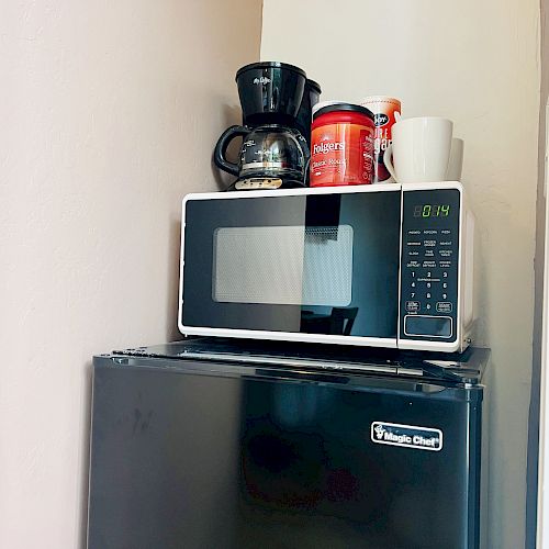 A small kitchen setup with a mini fridge, microwave, coffee maker, canister, and two mugs placed on a counter against a beige wall.