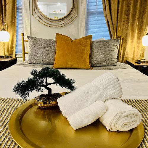 A neatly made bed with pillows, a tray holding rolled towels and a small potted plant, and ambient lighting from two bedside lamps.