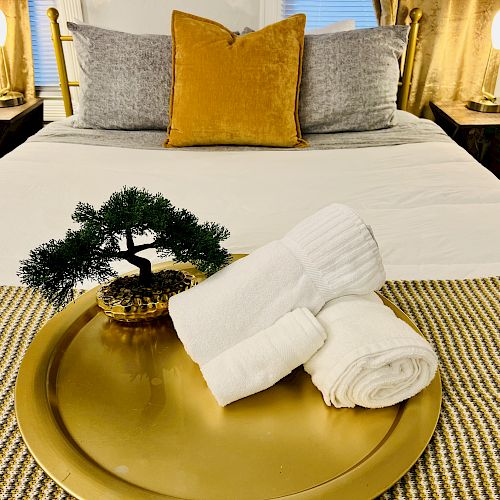 A neatly made bed with gray and gold pillows, flanked by lamps, and a gold tray with rolled white towels and a small decorative tree is in the forefront.