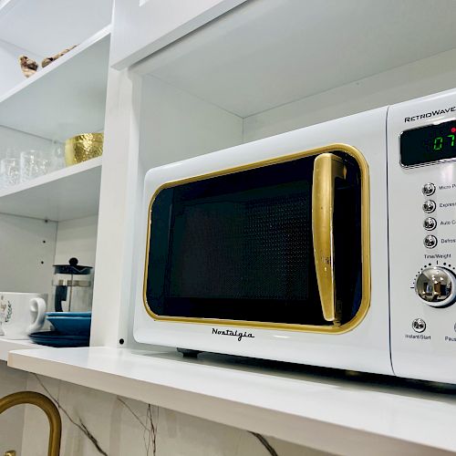 A white microwave with gold accents sits on a shelf in a kitchen with glassware and a French press nearby. The display shows 