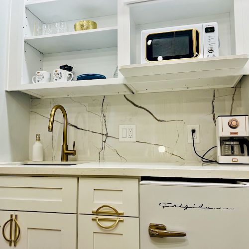 The image shows a small kitchenette with a sink, golden faucet, microwave, coffee maker, small fridge, and shelves holding cups and plates.