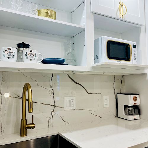 A modern kitchen with white cabinets, gold faucet, coffee maker, microwave, and open shelves with mugs and dishes.