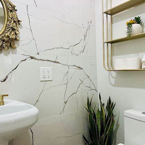 A bathroom features a white sink with gold fixtures, a round ornate mirror, marble wall tiles, a toilet, potted plant, and shelves with towels and decor items.