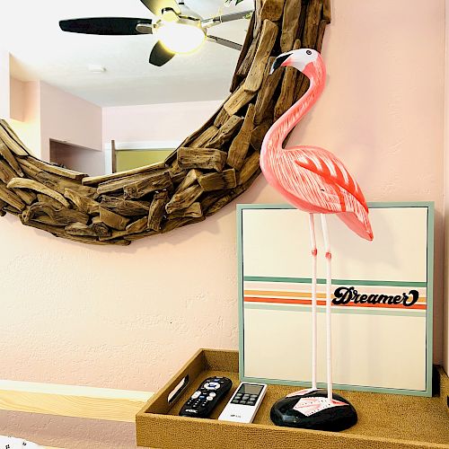 A pink flamingo figure, a “Dreamer” sign, a mirror with a rustic frame, a ceiling fan, and a remote control are placed on a wooden tray.