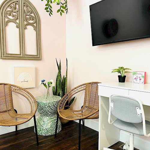 This image shows a cozy room with two wicker chairs, a small green table, a potted plant, and a white desk with a chair, featuring a wall-mounted TV.