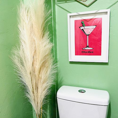 A green-walled bathroom with a white toilet, a framed cocktail art print, and a large pampas grass decoration next to it.