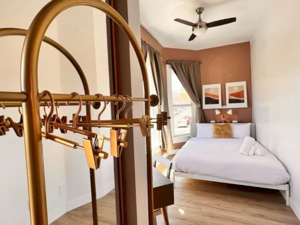 The image shows a modern bedroom with a neatly made bed, two wall paintings, a ceiling fan, and a luggage rack in the foreground.