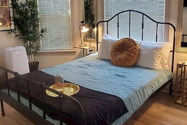 A cozy bedroom with a bed, blue sheets, decorative pillows, a tray, a lamp, plants, a mirror, wall art, a chair, and blinds on the windows.