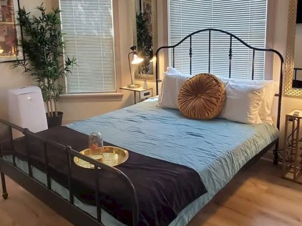 A cozy bedroom with a bed, blue sheets, decorative pillows, a tray, a lamp, plants, a mirror, wall art, a chair, and blinds on the windows.