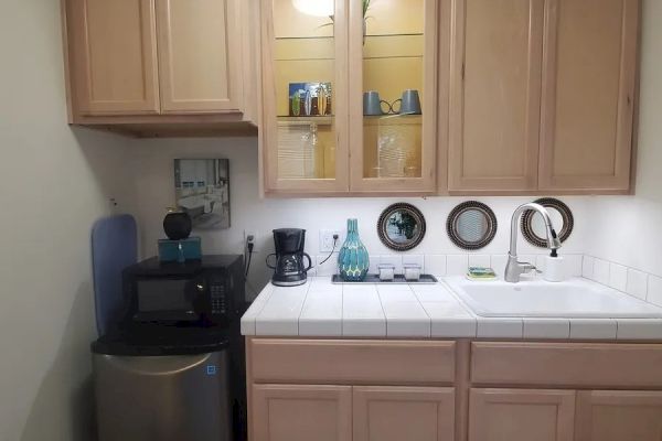 The image shows a small kitchenette with wooden cabinets, a countertop with a sink, a small refrigerator, a coffee maker, and a few kitchen items.