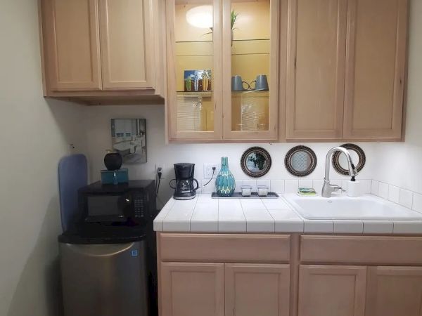 The image shows a small kitchenette with wooden cabinets, a countertop with a sink, a small refrigerator, a coffee maker, and a few kitchen items.