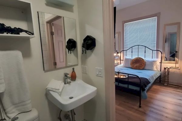 The image shows a bathroom with a sink and mirror, leading into a bedroom with a bed, large window, and wooden floor.