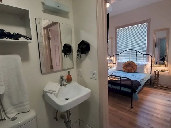 The image shows a bathroom with a sink and mirror, leading into a bedroom with a bed, large window, and wooden floor.