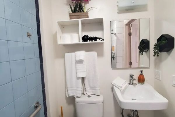 A small bathroom with a toilet, sink, mirror, white towels, bathroom shelves, a trash bin, and a flower vase on the top shelf for decoration.