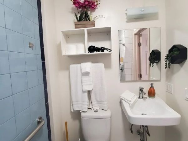 A small bathroom with a toilet, sink, mirror, white towels, bathroom shelves, a trash bin, and a flower vase on the top shelf for decoration.