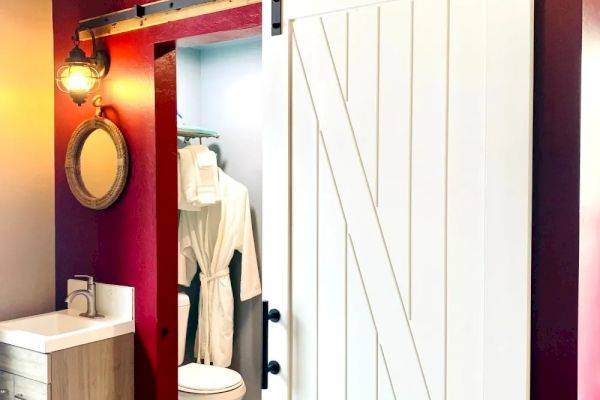 The image shows a bathroom with red walls, a white sliding barn door, a sink with a round mirror, and a visible toilet and hanging robe in the background.