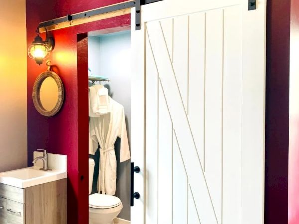 The image shows a bathroom with red walls, a white sliding barn door, a sink with a round mirror, and a visible toilet and hanging robe in the background.