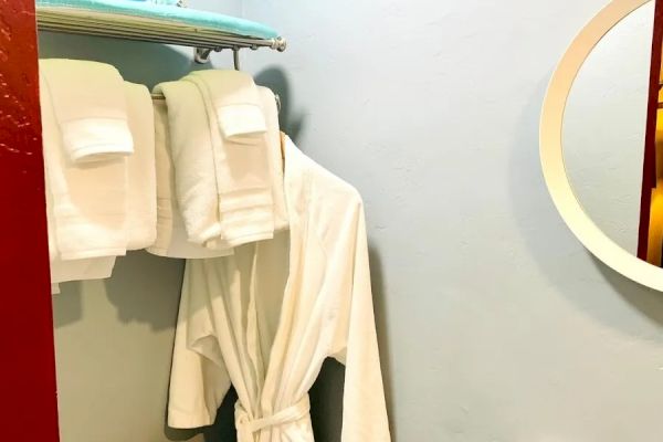 The image shows a small bathroom or dressing room with a bathrobe hanging, folded towels on a rack, a toilet paper holder, an iron, and an ironing board.