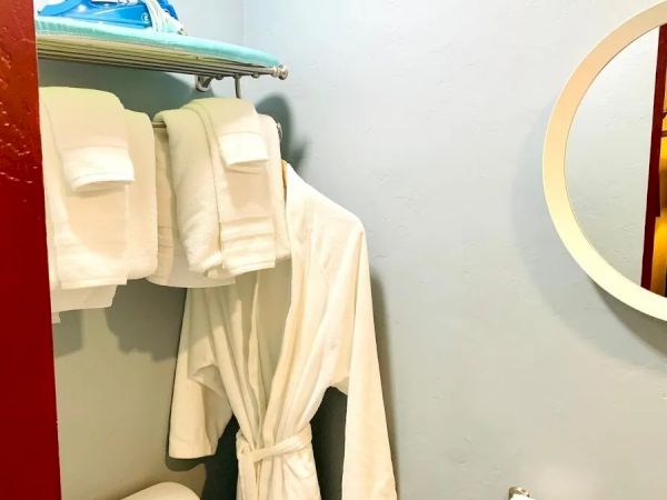 The image shows a small bathroom or dressing room with a bathrobe hanging, folded towels on a rack, a toilet paper holder, an iron, and an ironing board.