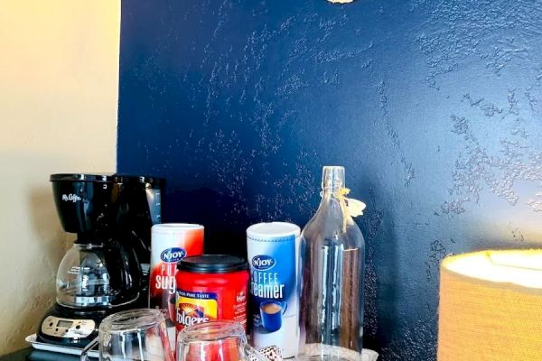 A coffee station with a coffee maker, mugs, coffee canisters, a bottle, and a seashell decor on a navy blue wall, next to a yellow lamp.