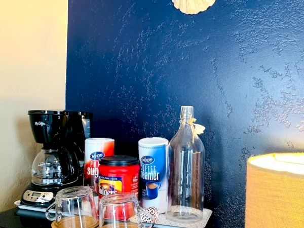 A coffee station with a coffee maker, mugs, coffee canisters, a bottle, and a seashell decor on a navy blue wall, next to a yellow lamp.