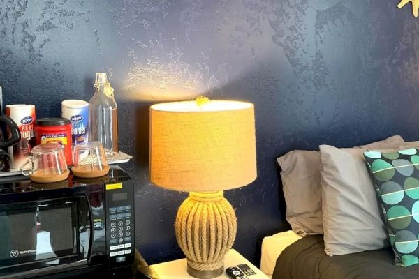 A cozy bedroom corner with a bed, nightstand, lamp, microwave, and some decorative items on the wall, including shells and a starfish.