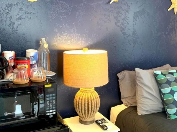 A cozy bedroom corner with a bed, nightstand, lamp, microwave, and some decorative items on the wall, including shells and a starfish.