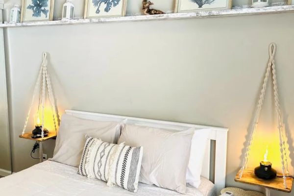 A cozy bedroom with a white bed, neutral pillows, hanging shelves with framed prints, and two side tables holding lamps and candles.