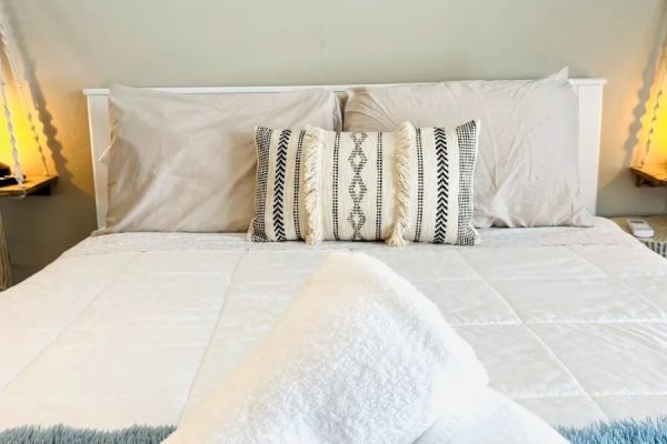 A neatly made bed with patterned pillows, rolled towels in the foreground, and soft lighting on either side of the headboard ends the sentence.