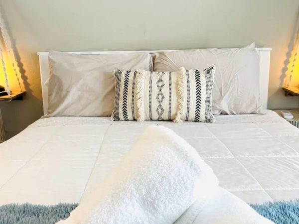 A neatly made bed with patterned pillows, rolled towels in the foreground, and soft lighting on either side of the headboard ends the sentence.