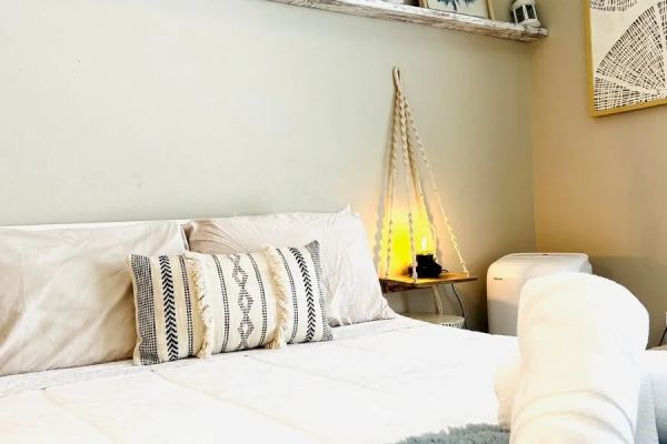 A cozy bedroom with a neatly arranged bed, decorative pillows, framed art on the shelf, a woven lamp, and a rolled towel at the foot of the bed.