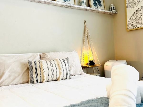 A cozy bedroom with a neatly arranged bed, decorative pillows, framed art on the shelf, a woven lamp, and a rolled towel at the foot of the bed.